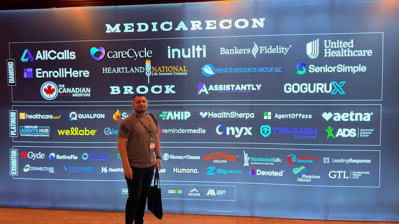 Wali Zuberi standing in front of the large MedicareCon 2026 backdrop at Ross Bridge Resort